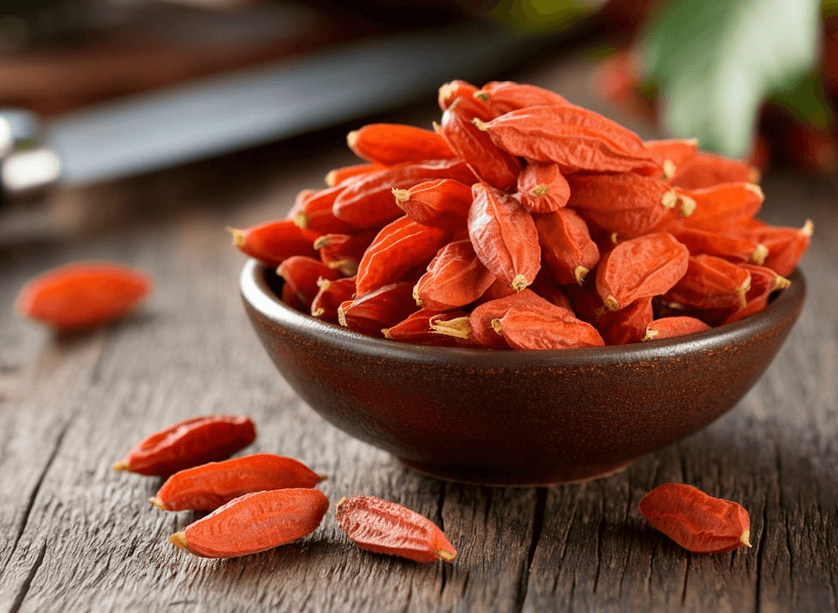 A bowl of goji berries with a few berries spilled on the table. The berries have a deep red color and a slight glossy sheen.